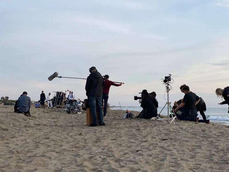 Professional location sound crew in Italy operating on a beach film set - A boom operator holding a long microphone boom pole with a windshield to capture dialogue, with camera operators and production crew setup on the sand during a seaside shoot.