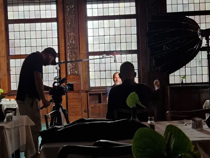 Behind-the-scenes of a professional ENG crew setup for a television interview shoot in Italy, featuring a camera operator on a tripod, an overhead boom microphone for location sound, and a studio LED light with a softbox diffuser.