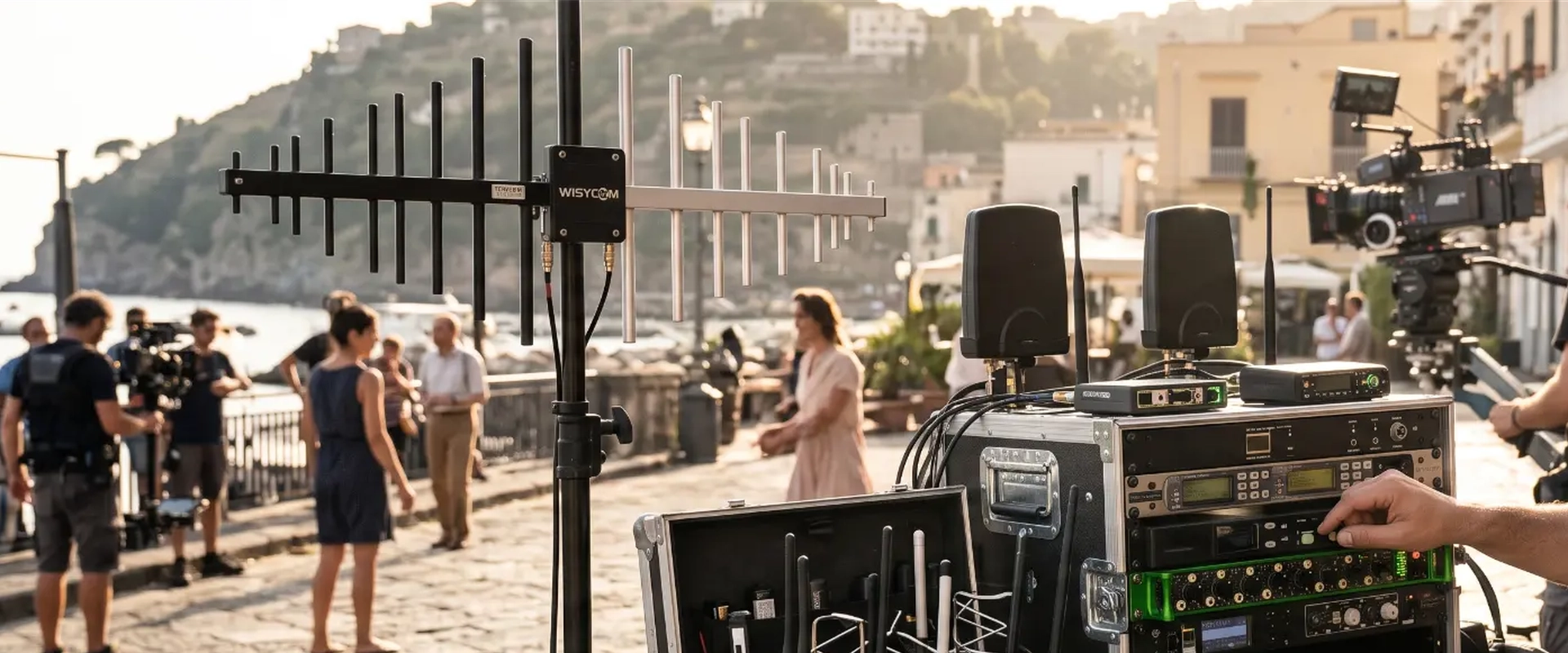Professional production sound mixer adjusting a wireless audio rack on an outdoor film set in Italy, featuring a large Wisycom directional shark fin antenna and RF splitter system mounted on a stand next to a heavy-duty flight case with wireless microphone antennas.